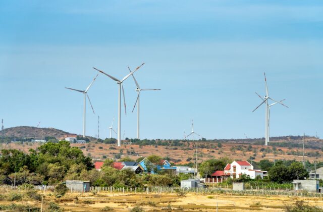 Small residential wind turbine on tall monopole in a rural backyard with neighboring houses visible in the distance
